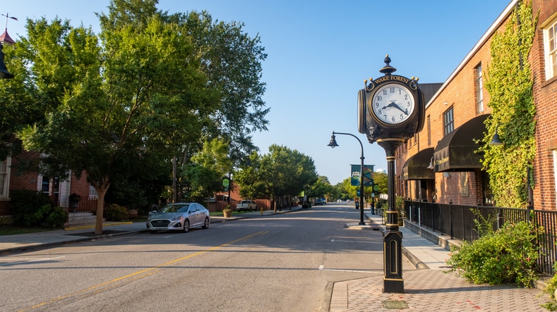 A quiet morning view of historic downtown Wake Forest, North Carolina, featuring classic brick storefronts, tree-lined sidewalks, and vintage street lamps under clear blue skies. The small-town atmosphere and warm sunlight highlight the charm and character of this Southern community.